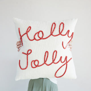Styled view of square natural cotton pillow with red Holly Jolly script embroidery