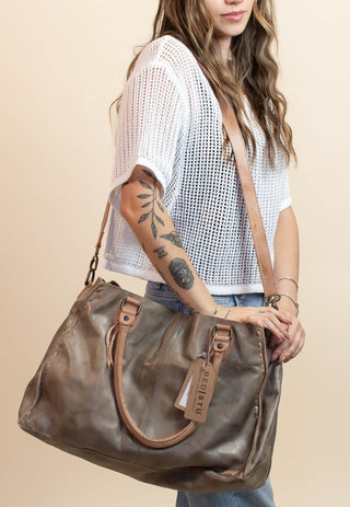 A woman modeling a large brown leather shoulder bag with a wide strap, standing against a solid beige studio background.