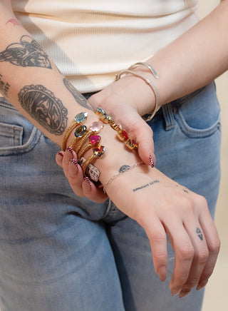 A close-up of a woman's tattooed arm wearing a stack of colorful beaded bracelets, a gold chain bracelet, and a silver bangle, paired with a cream ribbed tank top and light blue jeans.
