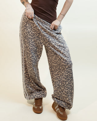 A woman modeling a pair of loose, wide-leg leopard-print pants with a brown ribbed tank top and brown shoes, standing against a plain cream studio background.
