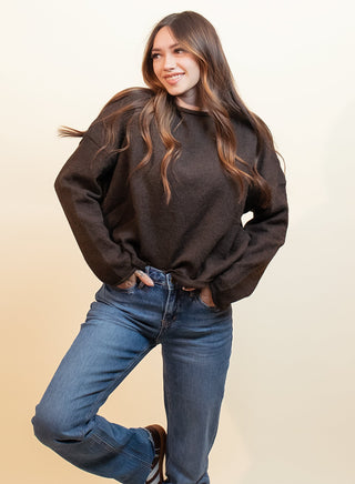 A woman posing in a dark brown crewneck sweatshirt, blue jeans, and brown suede shoes against a warm beige studio background.