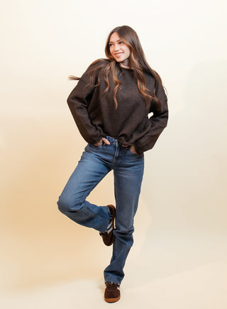 A woman posing in a dark brown crewneck sweatshirt, blue jeans, and brown suede shoes against a warm beige studio background.