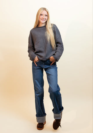 A woman with long blonde hair stands against a warm beige studio background, wearing a dark gray crewneck sweater, blue jeans with cuffed hems, and dark brown boots.