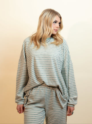 A blonde woman wearing a matching two-piece lounge set with a sage green and cream horizontal stripe pattern, standing against a warm beige studio background.