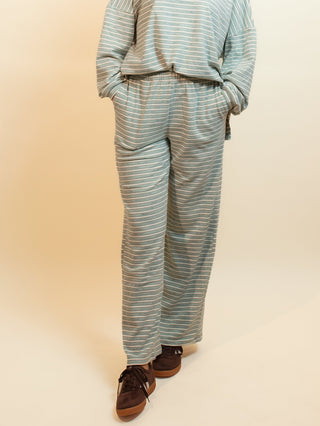 A woman modeling a matching two-piece loungewear set consisting of a long-sleeve mock neck top and wide-leg pants, both in a light blue and white striped pattern. She is standing against a warm beige studio backdrop.