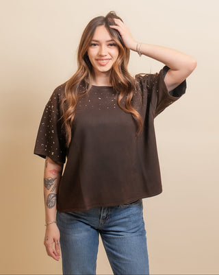 A woman modeling a brown short-sleeve top with sheer, embellished sleeves, paired with blue jeans, against a solid beige background.