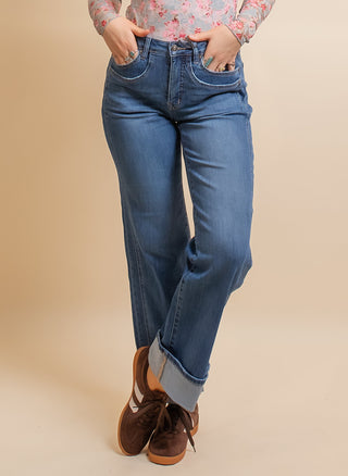 A woman modeling a sheer long-sleeve mock neck top with a pink and blue floral pattern, paired with blue jeans and brown shoes, standing against a solid beige studio background.