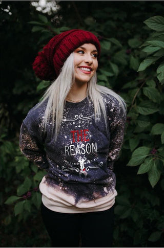 A woman wearing a dark tie-dye graphic sweatshirt with 'THE REASON' printed on the front, a dark red knit beanie, and a cream undershirt, standing outdoors in front of a background of green foliage.