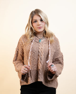 A woman modeling a chunky, cable-knit cropped sweater in a warm taupe-brown color, paired with black pants and layered necklaces, set against a solid light beige background.