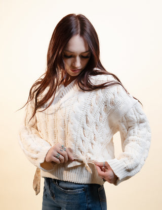 A woman with long dark brown hair wearing a cream cable-knit sweater and blue jeans, looking down as she adjusts the tie at the waist of her sweater against a solid light beige background.