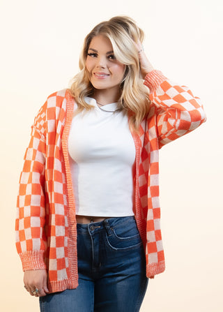 A woman modeling a bright orange and white checkered cardigan over a white crop top, blue jeans, and black sneakers, standing against a solid beige studio background.