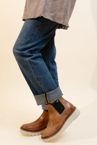 A person wearing a taupe knit cardigan, blue jeans with rolled cuffs, and brown leather Chelsea-style boots with white soles, standing against a plain cream background.